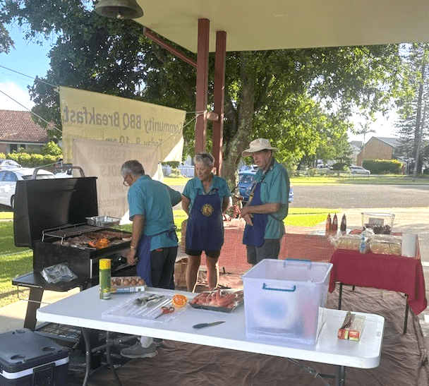 Three Lions members in aprons over blue shirts beside food-covered tables and a BBQ full of cooking food.